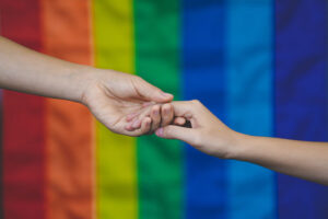 two individuals hold hands against a rainbow flag backdrop