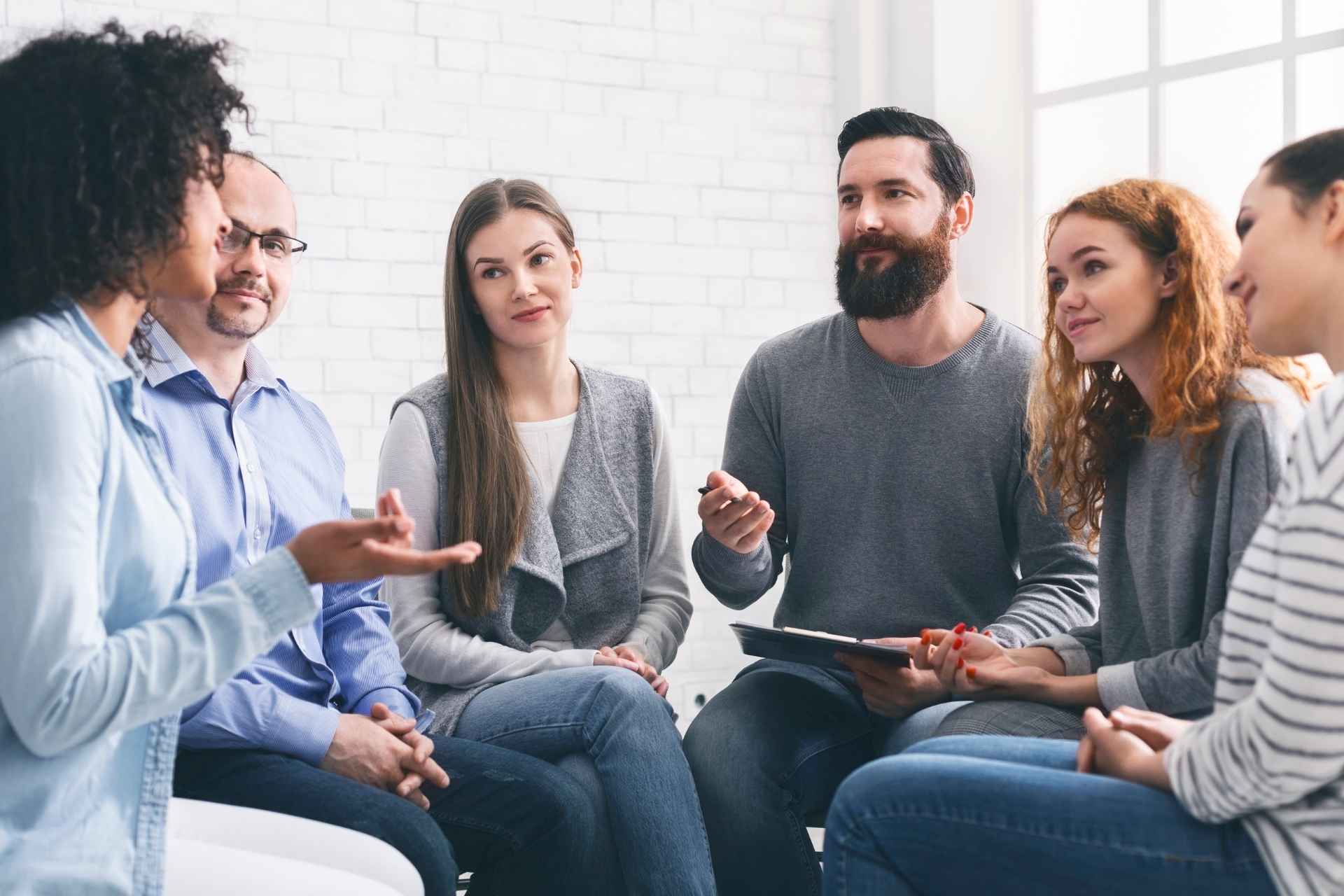 Six adults sitting in a circle and participating in a group discussion, with one woman speaking while others listen.
